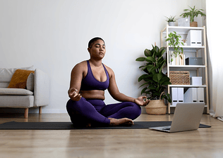 A woman meditating on a yoga mat in a cross-legged position. Her eyes are closed and her hands are resting on her knees. She has a laptop open in front of her that could be playing a guided meditation for self-confidence.
