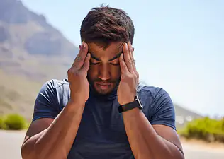 A close-up photo of an athlete outdoors. He is wearing a blue shirt and has his hands on his temples with his eyes closed. He is working out with a headache.