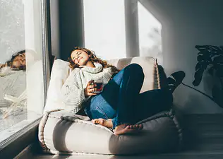 A woman practicing self-care. She's resting on a comfy chair, basking in the sun, and drinking tea.