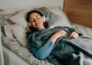A relaxed woman meditating in bed. She has headphones on and is under a blanket.