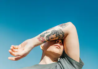 A person covering their eyes with their tattooed arm. They are feeling sick from stress. The background is a bright blue sky.