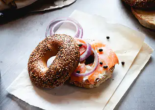 An everything bagel with cream cheese, salmon, red onions, and capers resting on a brown parchment bag.