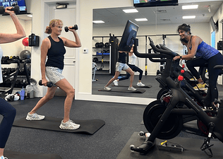 One person riding on a Peloton Bike+ on the right, and two people lifting dumbbells on mats on the left, taking a class on Peloton Bike+ with the screens turned in The Moorings Club fitness center.