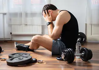 A man dealing with exercise burnout. His head is in his hands and he's feeling mentally drained, surrounded by dumbbells and weights.