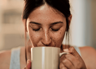 Does Coffee Dehydrate You?: Close-up photo of a woman with freckles drinking a cup of coffee from a mug.