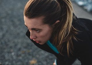 A young woman with a ponytail is bent over outside with her hands on her knees, experiencing nausea after a workout.