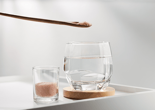 A tiny wooden spoon of pink Himalayan salt hovering over a glass of drinking water. There's a small jar of salt next to the glass as well.