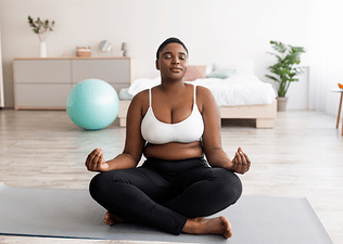 A woman sitting on a yoga mat at home doing a postpartum workout. She is in lotus position with her eyes closed.