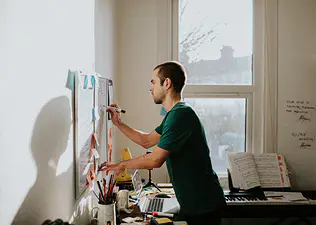 A man leaning over his desk and writing on a whiteboard in a sunny home office room. Learn "how long does it take to form a habit?" in this article.