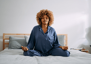 A woman practicing daily meditation while sitting on her bed in a sunny bedroom.