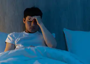A man experiencing anxiety at night. He's sitting up in bed, closing his eyes, and rubbing his forehead in stress.