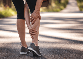 A close-up photo of a woman grabbing her cramping calf muscle during an outdoor run.
