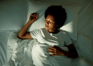 A woman practicing sleep meditation in bed. She's lying on her back under the covers with her eyes closed.