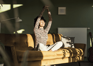 A woman stretching her arms overhead while closing her eyes and smiling. She is sitting on the couch in the sunlight.