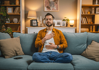  A man sitting on the couch at home with his hands on his chest and belly, practicing a loving-kindness meditation.