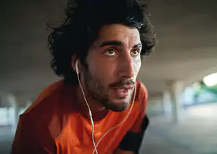 A close-up photo of a sweaty young man listening to headphones who just finished his workout.