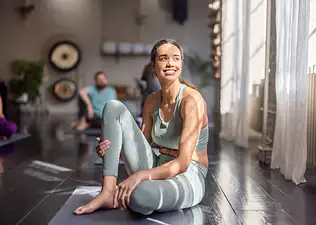 A woman sitting down on a yoga mat after exercising, smiling and looking away.