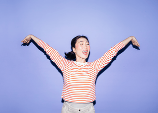 A woman smiling and spreading out her arms energetically. She's wearing a red and white striped shirt and standing against a lavender background. Learn how to get more energy in this article.