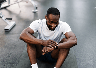 A man sitting down at the gym, smiling and looking at his smart watch.