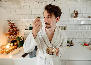 A man in his bathrobe eating oatmeal and fruit in his kitchen.