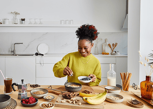 A happy woman at home preparing a healthy breakfast with complex carbohydrates like fruits and nuts.