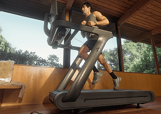 Man running on a Peloton Tread treadmill inside a room with large glass windows and wooden ceiling.