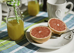 A plate with a grapefruit cut in half next to a bottle of fresh orange juice, both of which are hydrating foods and drinks.