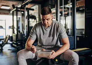 A man eating carbs in order to build muscle. He's sitting on a workout bench eating oats from a plastic container.