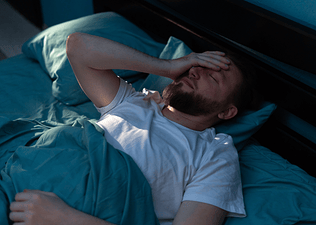 A man experiencing sleep anxiety. He's lying in bed at night with his hand wearily over his forehead.