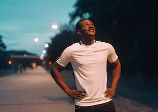 A man recovering from an outdoor walk or jog at night while looking up at the sky and putting his hands on his hips. 