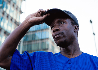 A sweaty man adjusting his baseball hat while he stands outside and looks into the distance.