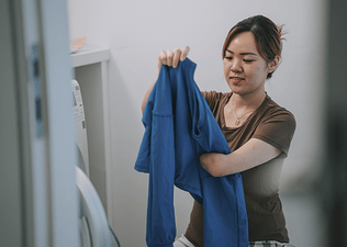 A woman holding up and folding a blue long-sleeve shirt at home in the laundry room.