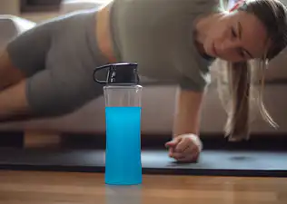 A blue sports drink in the foreground with a woman doing a side plank on a workout mat in the background.