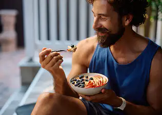 A man eating a bowl of oats topped with fresh fruit while sitting outside. He's eating carbs before his workout.