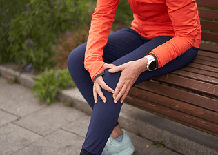 A woman experiencing runner's knee. It's a close-up photo of her sitting on a park bench after a run and holding onto the front of her left knee.