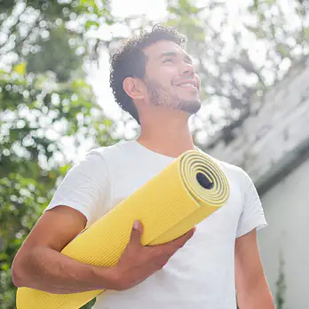 A man holding a yellow yoga mat and smiling while standing outside. Learn how to motivate yourself to work out here.
