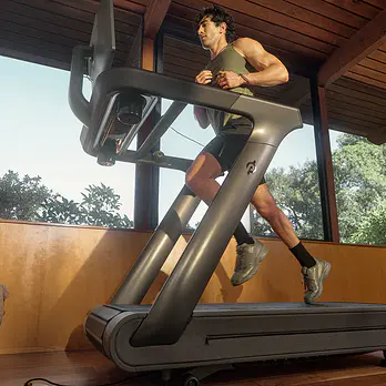 Man running on a Peloton Tread treadmill inside a room with large glass windows and wooden ceiling.