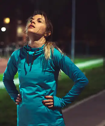 An exasperated athlete on an outdoor running path in the evening, with her hands on her hips and head pointing up to the sky. She has lost motivation to work out.