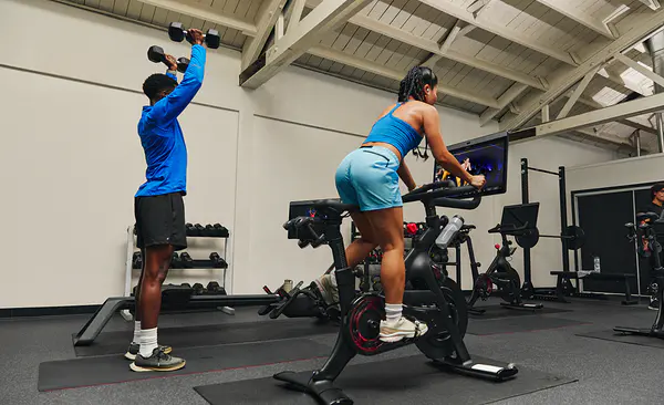 Two friends doing strength and cardio workouts side by side in a gym using a Peloton Bike.