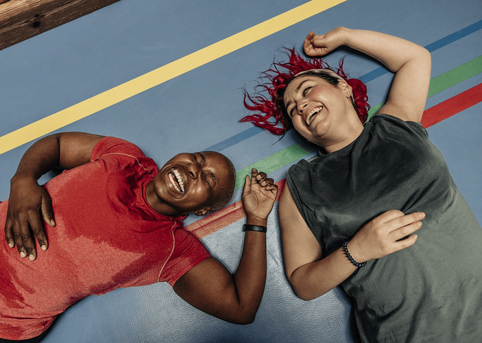 Two women lying down on a colorful exercise mat after exercising, laughing. Exercising is a great way to activate your "happy hormones."
