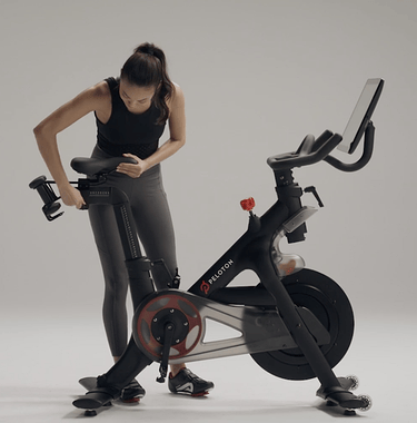 Woman setting up her Peloton Bike, adjusting the handlebars and seat height, clipping into the Bike pedals, and showing different riding positions.