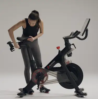 Frau richtet ihr Peloton Bike ein, stellt den Lenker und die Sitzhöhe ein, klickt sich in die Bike-Pedale und zeigt verschiedene Sitzpositionen.