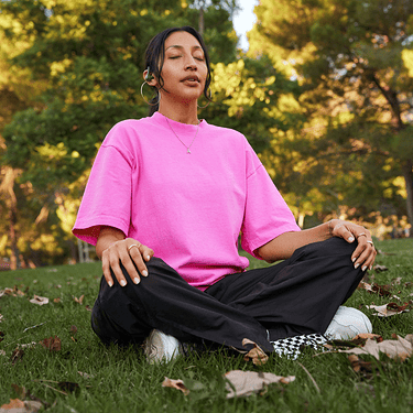 A young person sits cross legged on grass surrounded by trees with earphones in and a smartphone at their feet.