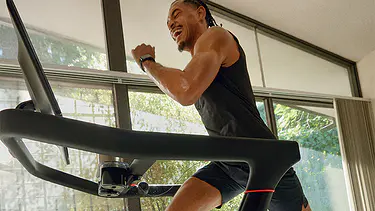 A happy smiling man runs on a Peloton Treadmill in a well light room with windows opening to outdoors with scenes of greenery