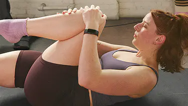 A lady wearing maroon and purple fitness shorts and top stretching her knee to chest working out with the Peloton App