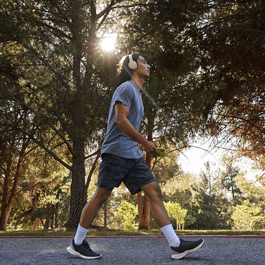 A young person walks outside in a wooded area while wearing headphones and holding a smartphone.