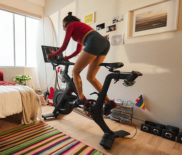 A woman wearing Peloton apparel pedals a stationary Peloton bike in a sunny apartment living room