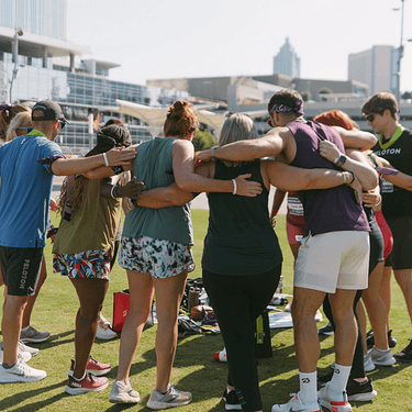 A group of people in workout clothes huddle in a circle while standing outside on a turf field.