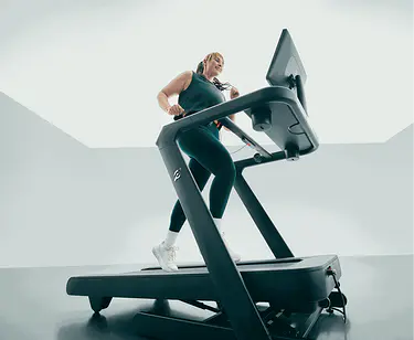 A smiling woman running on a Peloton treadmill in a brightly lit room.