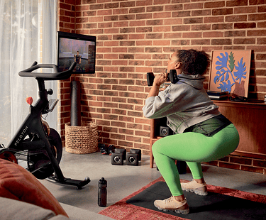 A woman in workout clothes does squats with dumbbells in front of a Peloton bike displaying a live class